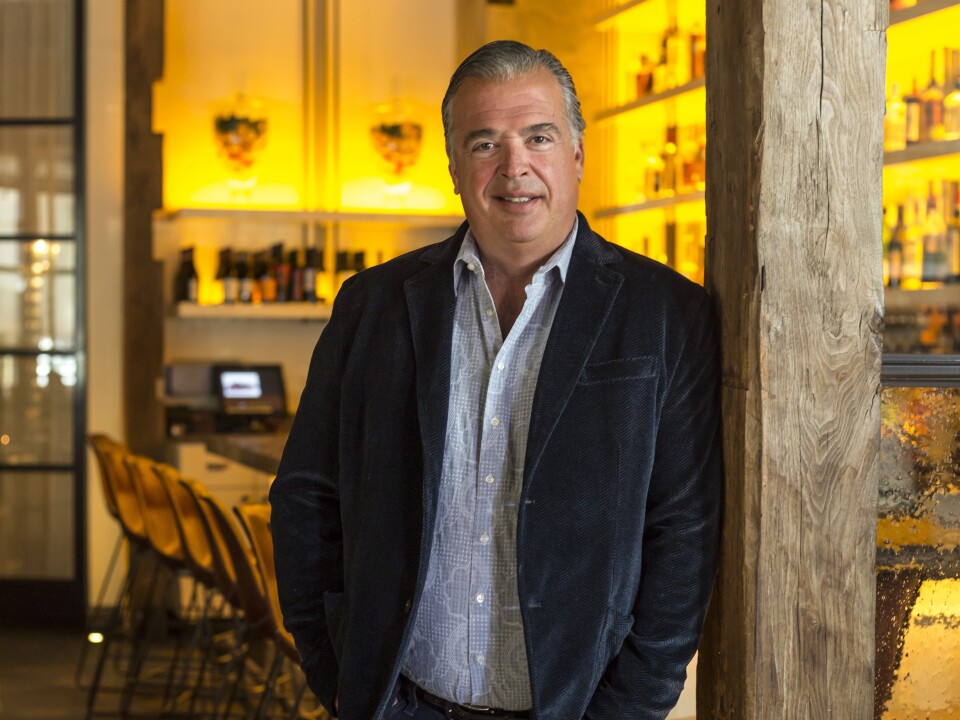 A man with light-tone skim leans on a wood pole in a restaurant bar. He wears a jacket over an open-collar shirt.