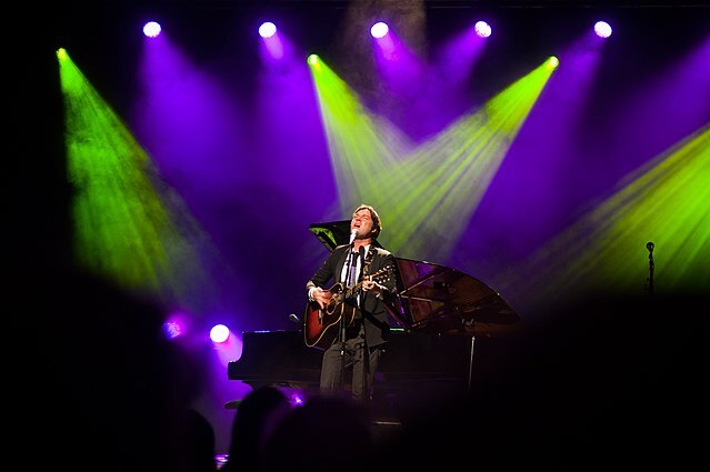 Rufus Wainwright playing guitar on stage under green and purple lighting.