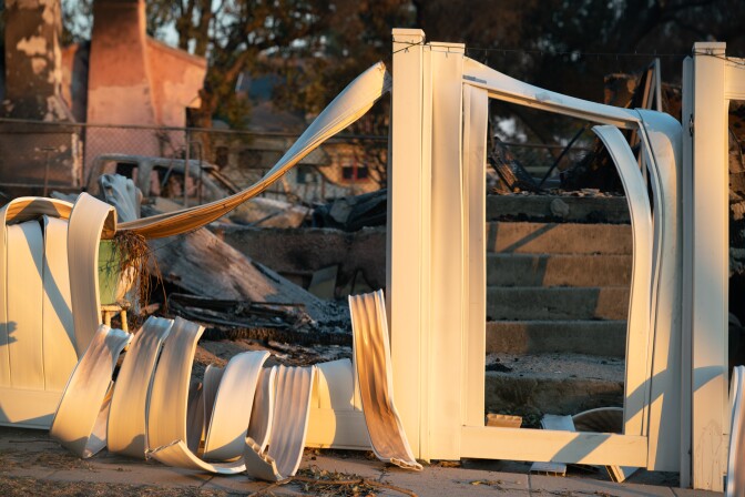 A white melted fence is melted in front of a destroyed home where remains of chimneys and cars remain.