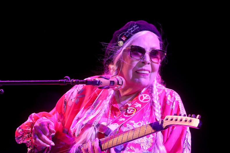 Joni Mitchell, dressed in pink an bathed in pink light, holds a guitar on stage with a microphone in front of her as she performs.