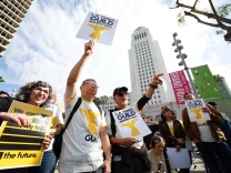 A group of people, some wearing white shirts with a yellow eagle and black text that reads "Guild" gather outdoors holding white, yellow, and black signs that reads "L.A. Times Guild Against ULPs." Los Angeles City Hall is in the background. 