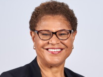 Headshot of Karen Bass. She has light brown skin and short, curly light brown hair. She is smiling and wearing a dark suit jacket or blouse, black and brown horn-rimmed glasses, and small hoop earrings.