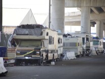 Four tan and white RVs are parked along an underpass. Yellow crime scene tape is attached to the closest RV, and it's covered in tarps and debris with a wooden board blocking out the front windshield. The vehicles look like they haven't moved in a while, if they can move at all.