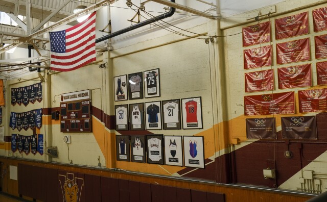 An American flag hands from the rafter of a high school gym. The primary colors on the walls and banners are burgundy and golden yellow. On the right, more than a dozen identical banners with the Olympic rings list the names of alumni that have competed in the Summer Games.