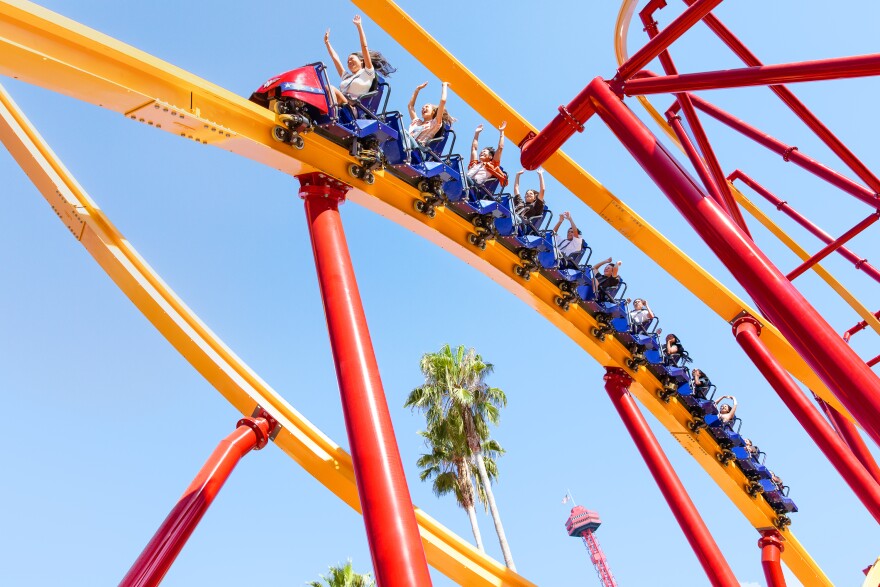 The Wonder Woman Flight of Courage roller coaster, with fans holding up their arms as they ride on the yellow tracks, with red scaffolding supporting them in their red and blue ride cars.