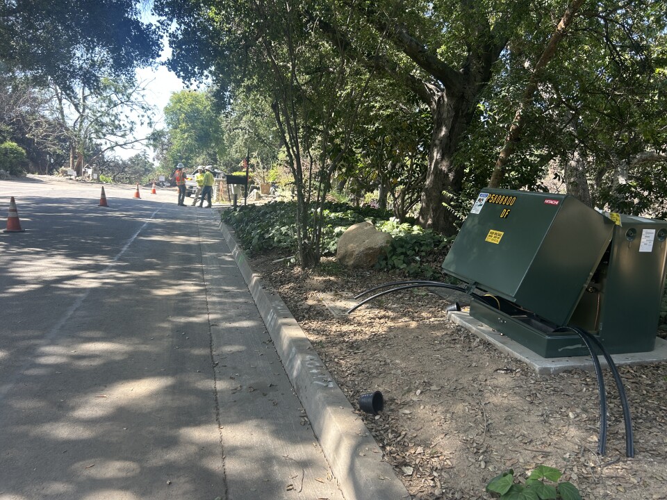 A green transformer box next to a newly paved street under the shade of oak trees. 