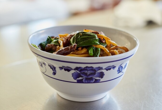 A small white bowl adorned with pictures of blue flowers sits on a metal countertop. The bowl contains a dish made up of noodles, slivers of meat and sauteed greens. 