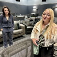 Two female presenting persons stand inside a room painted gray. Several couches are behind them.