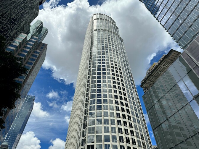 A curved skyscraper rises like a giant pillar against a vibrant blue sky, and it's capped by a billowy white cloud. It's surrounded by other tall steel and glass buildings.