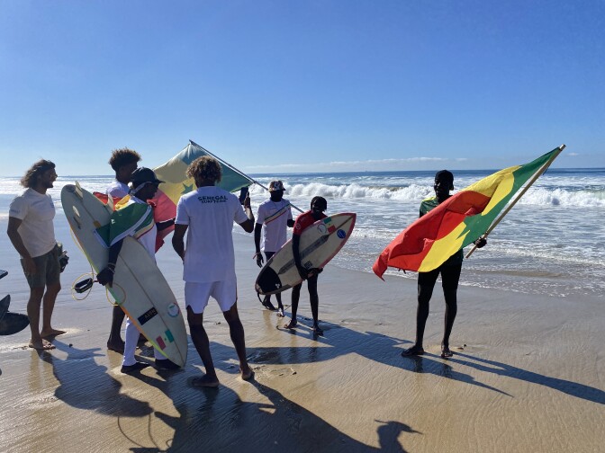 Six men in white t-shirts, two of them waving red, yellow and green Senegalese flags, greet a Black woman with short hair, wearing a red jersey and holding a surfboard on the beach. 