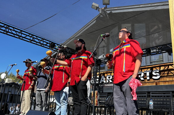 Performers in red shirts with blue and yellow trim shake maracas and sing into microphones on an outdoor stage.