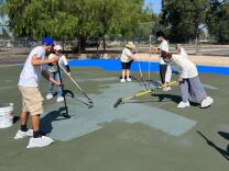 Half a dozen people are painting asphalt at a park using long paint rollers.