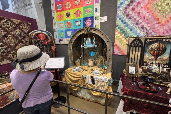 A woman in a purple t-shirt and gray straw bucket hat with black bow looks at table setting displays. In the center is an ocean-themed table with a blue octopus chandelier, gold draped tablecloth, and a porthole in the background.
