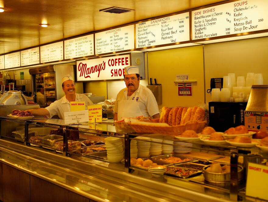 A pair of deli workers wearing aprons and white hats stand behind a counter, surrounded by classic deli dishes, with bright signs behind them showing the menu