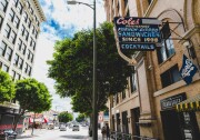 A sun-drenched downtown L.A. street scene with the iconic vintage neon sign of Cole’s—"Originators of the French Dipped Sandwiches Since 1908"—glowing proudly above the sidewalk. 