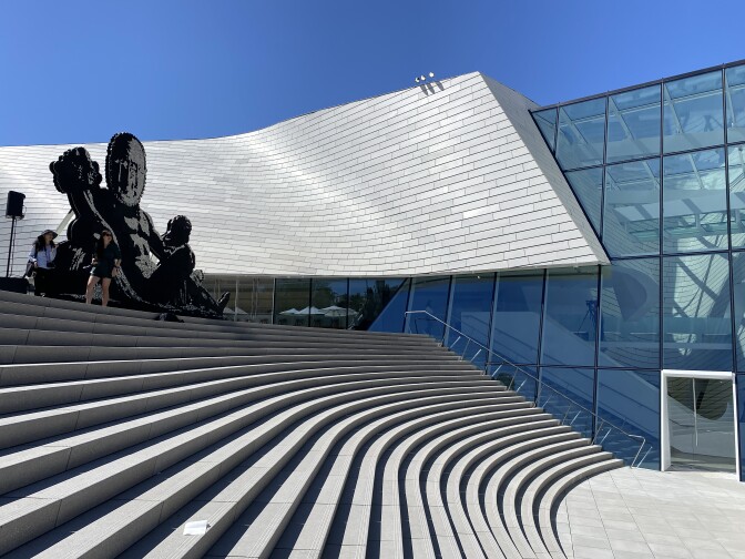 A wide outdoor staircase is in the foreground with the curved, silver-colored sloping roof of a building in the background. The sky is a clear bright blue behind the building.