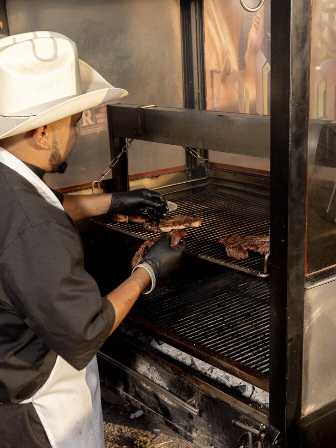 A cook with medium dark skin tone in a white cowboy hat and black gloves grilling strips of steak over a charcoal fire in a large metal grill.