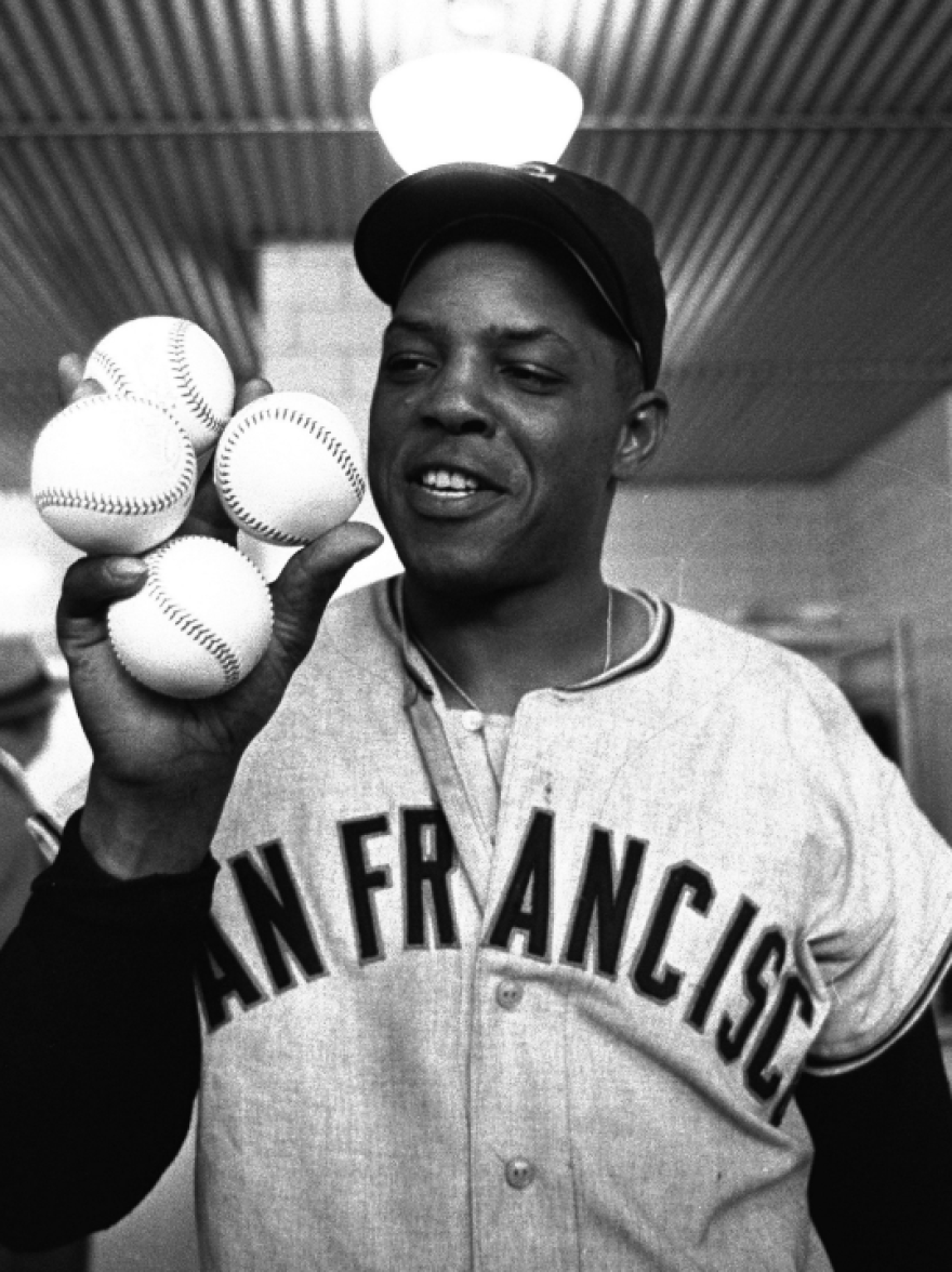 A man is holding up four baseballs in one hand. He's smiling and wearing a San Francisco jersey. 