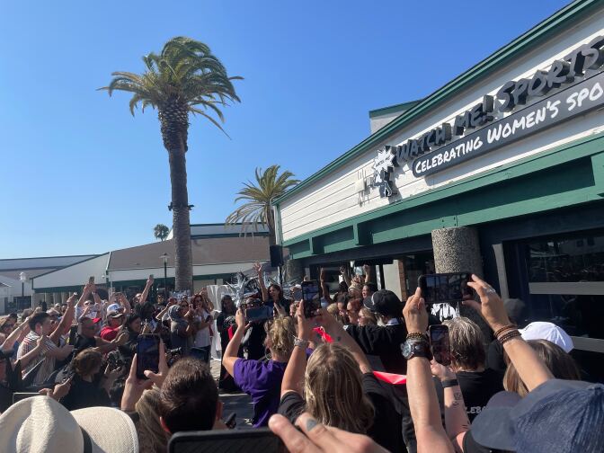 A crowd holds their phones up outside a bar. The words "Watch Me! Sports" and "Celebrating women's sports" are visible on the sign.
