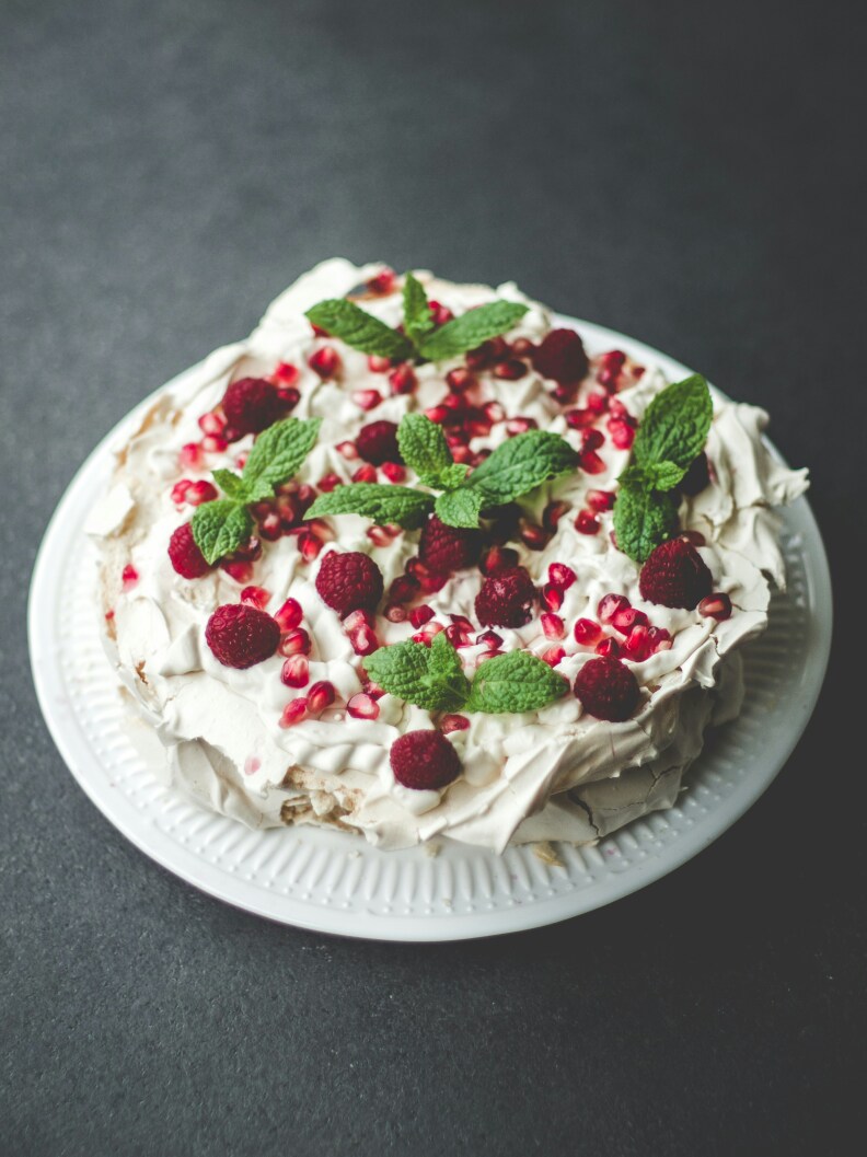 A dessert resembling a cake, made with whipped white meringue, featuring uneven edges and adorned with whole red raspberries, pomegranate seeds, and fresh green mint leaves.