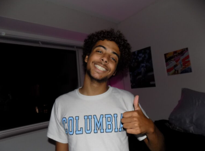 A Black man poses for a photo. He is smiling and has dark brown hair and light facial hair. He is giving a thumbs up and is wearing a grey or white shirt that says Columbia in light blue letters with a black outline.