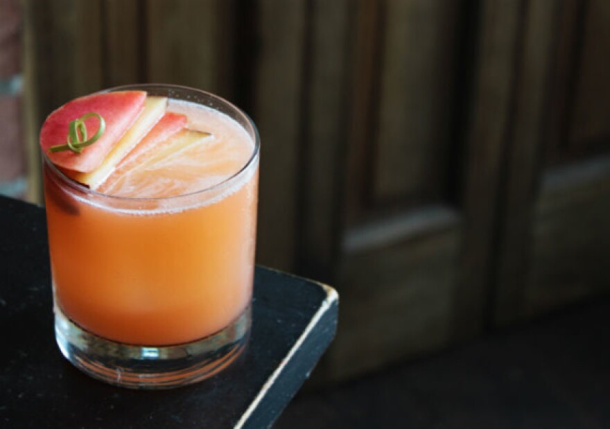 a peach-colored drink in an old fashioned glass sits on the edge of a black bar