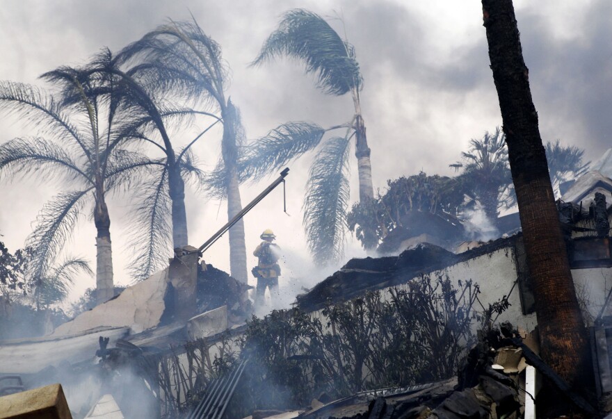 A firefighter stands under windswept palm trees as he hoses down smoldering debris in Ventura, Calif., Tuesday, Dec. 5, 2017. Ferocious Santa Ana winds raking Southern California whipped explosive wildfires Tuesday, prompting evacuation orders for thousands of homes.