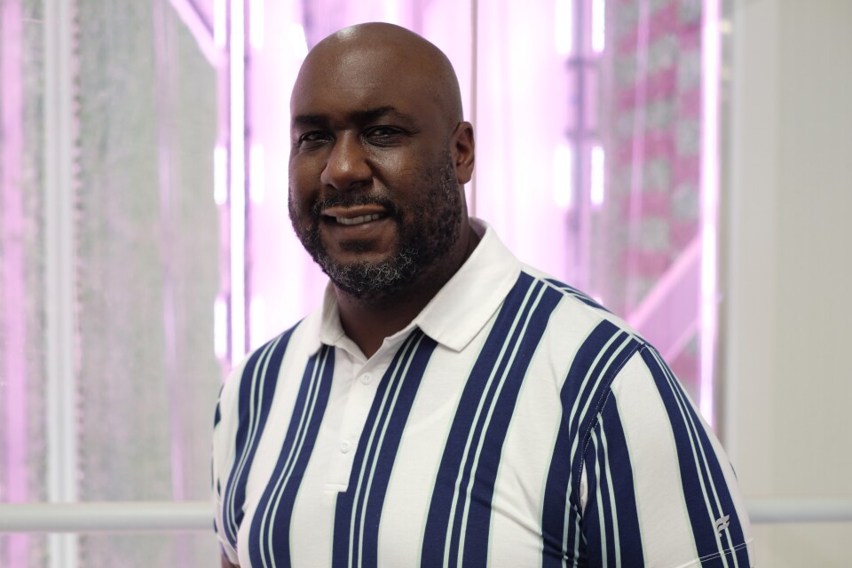 A Black man with a short salt and pepper beard and mustache wearing a white and blue vertically striped shirt smiles for the camera. The background is blurred white and purple. 