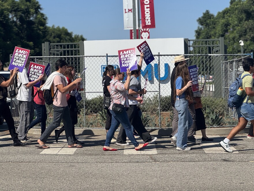 About a dozen, mostly dressed in denim, walk around a building area labeled LMU. They are holding up signs that read "LMU: Back to the table" and "Union busting is not a Jesuit value."