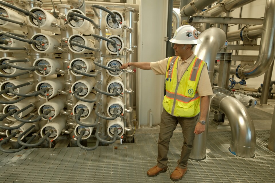 A man in a white hardhat and neon safety vest points to a machine that puts wastewater through reverse osmosis to purify it. 