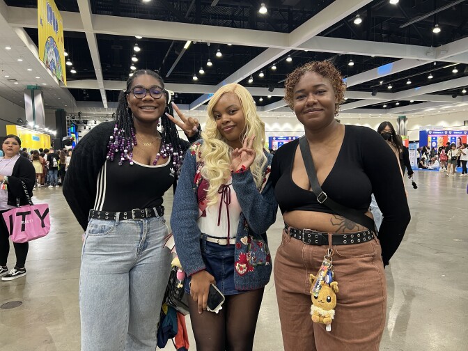 Three brown-skinned women stand in a photo together in a convention hall. 