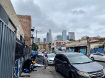 The LA skyline as seen from Skid Row. There are tents lining one side of the sidewalk next to parked cars.