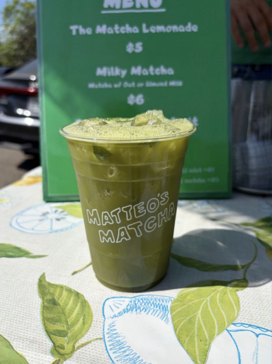 A green iced matcha drink in a cup reading 'Matteo's Matcha' sits in front of a menu board.