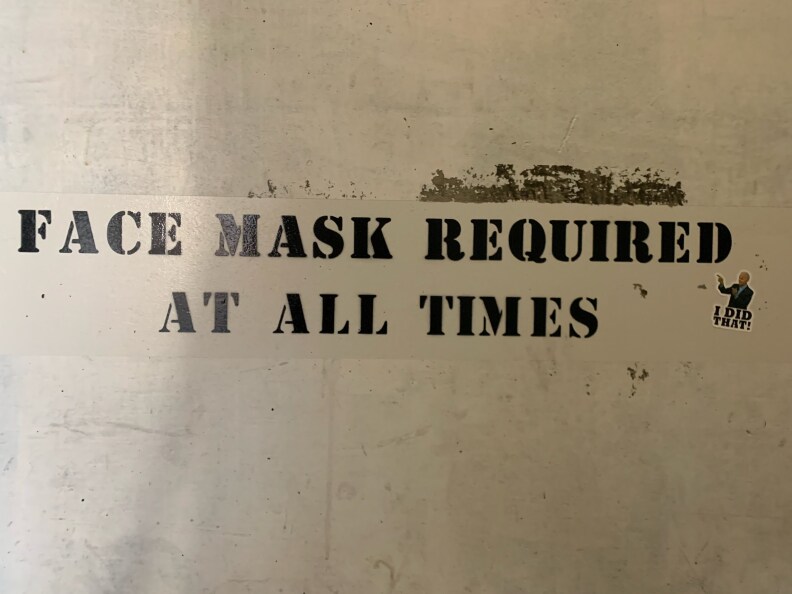 A sign stenciled in black on a parking garage wall reads, "Face mask required at all times." Just below the message is a small sticker that is a photograph of President Biden pointing to the sign’s message above the words, “I did that!”