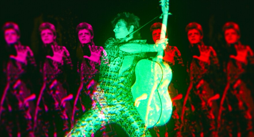 A kaleidoscopic image with a green David Bowie holding a guitar in the center, and purple and red Bowies to each side.