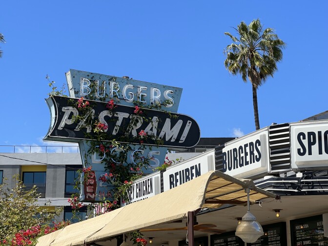 A mid-century modern restaurant sign advertising burgers, pastrami and hot dogs sits atop the Johnny's West Adams building. The backdrop is a blue sky, a palm tree and a new office building.