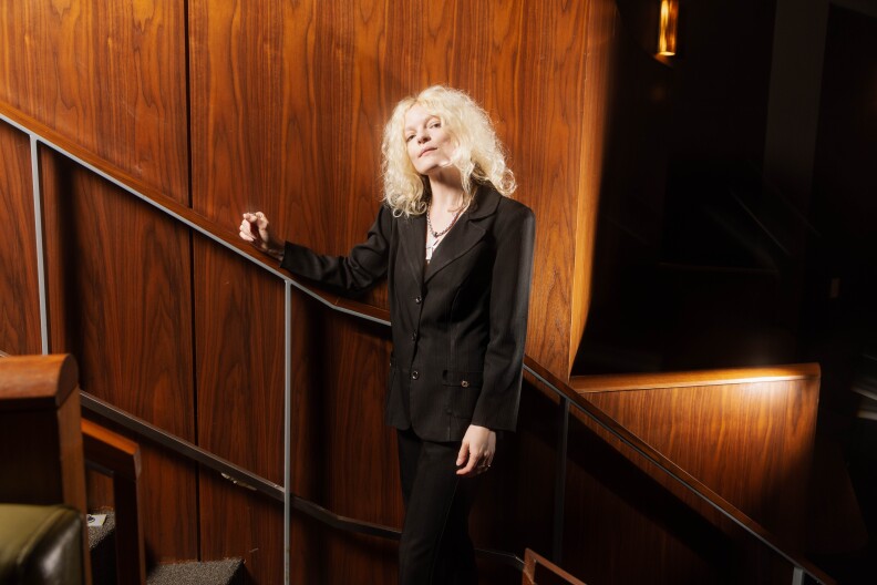 Jessica Pratt wears a black suit and has platinum blonde, should-length hair. She's posted on a staircase, standing a looking to camera. Behind her is a wood-paneled wall.