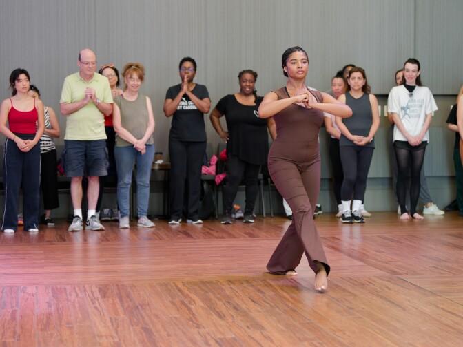 A woman in a brown jumpsuit performs a dance movement with her arms held in front of her chest and feet crossed on a wooden studio floor. A diverse group of participants stands in the background, observing and preparing to follow along.