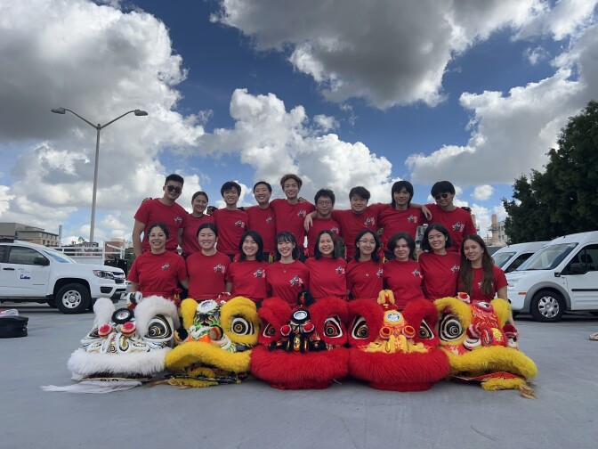 A group photo of 18 college students, all wearing red t-shirts and smiling. They are in two rows, the back row standing and the front row kneeling, and in the very front are five furry Chinese lion dance costume heads -- two red, two yellow and two white. They're posing in a parking lot with cars parked around them and the sky is blue with bright white clouds.