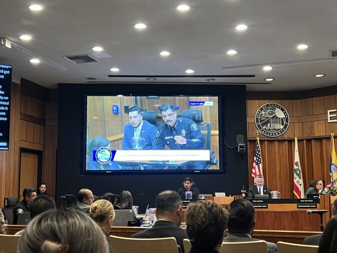 A crowded city council meeting room: Several elected officials are sitting on a dais, with flags hanging behind them. There is a screen showing two individuals speaking to the city council, one is wearing a police uniform.