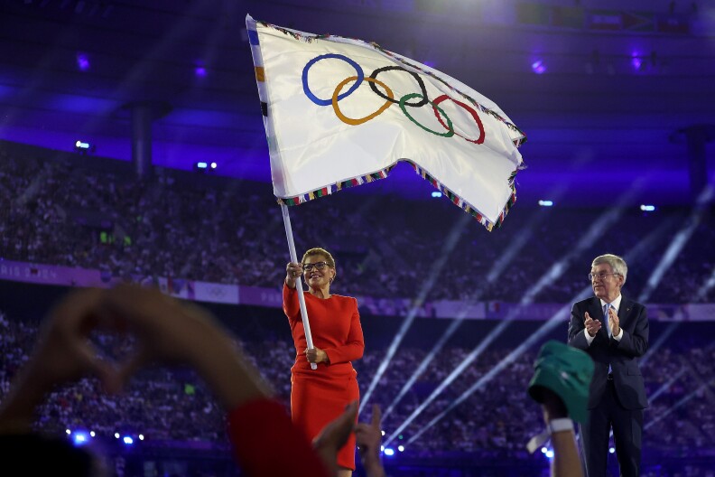 A Black woman in a red dress waves the Olympic flag as a white man in suit applauds.