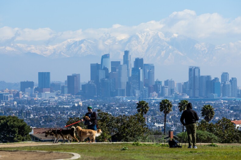 A view from a sunny park of the downtown Los Angeles skyline with snow-capped mountains in the background.
