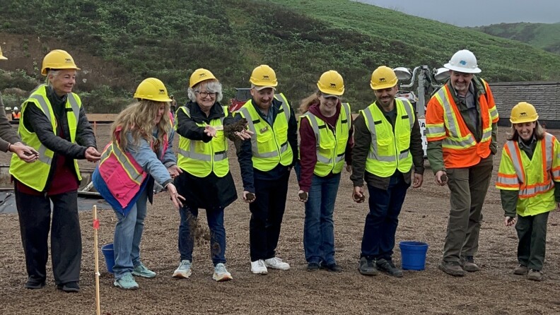 At least eight people, all wearing yellow hard hats and neon high-visibility vests, are throwing handfuls of dirt on the ground in front of them with big smiles on their faces.