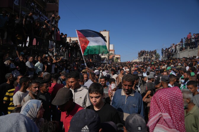 A large group of people gather in the street and stand on top of buildings. A person hoists a Palestinian flag. 
