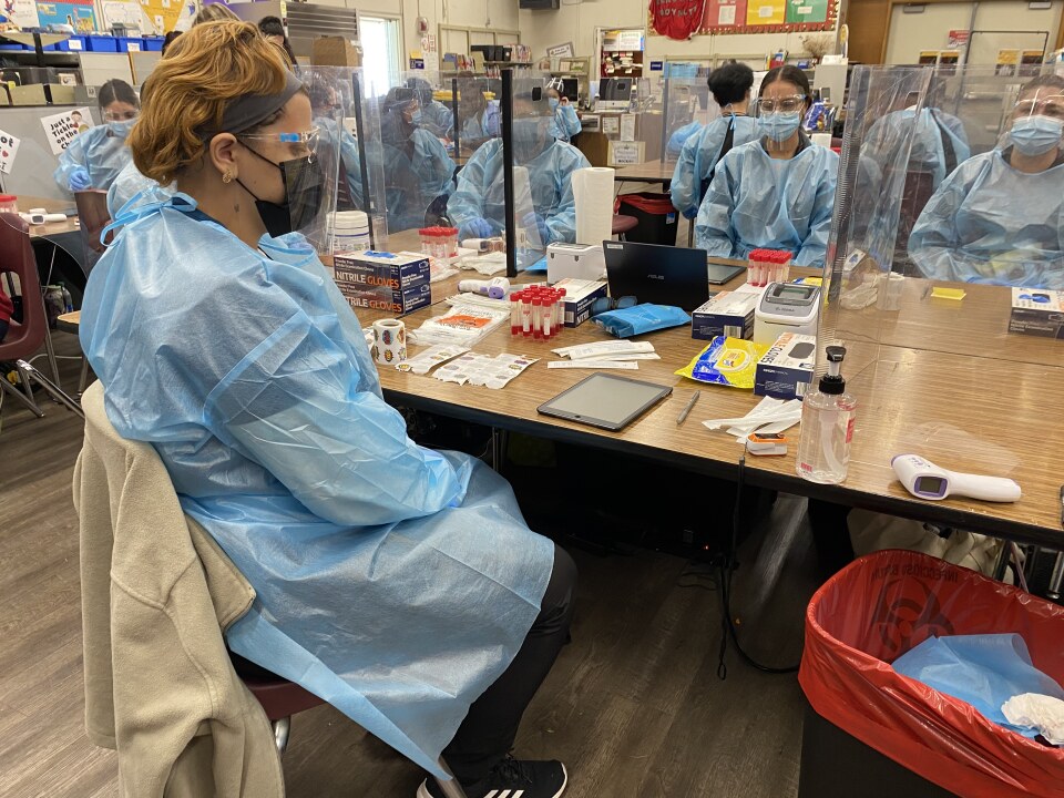 People sit at tables in front of vials, swabs and other testing equipment. The tables are divided by plastic dividers, with an adult wearing a blue plastic gown over their clothing sitting in each section. The people are also wearing masks over their noses and mouths and plastic protective glasses over their eyes. 