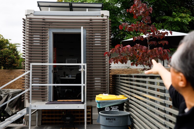 A man out of focus on the bottom right of frame points to a gray, vertically rectangular structure with a small stair case, slabs, and an open door.