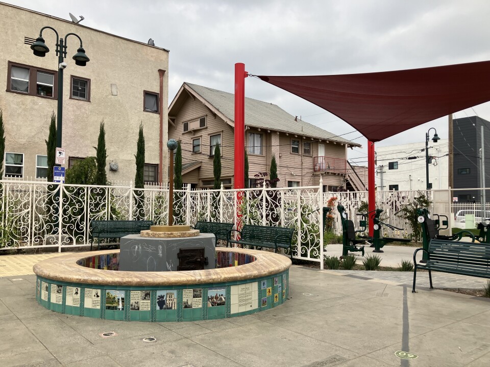An outdoor park scene: a park fountain covered in illustrated aquamarine blue tiles, with a white wrought iron fence behind it, and a beige apartment building and large pink house in the background.