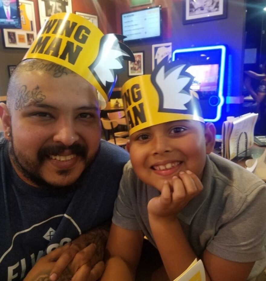 Brandon Lopez is smiling with his son, Sonny, next to him. They both wear yellow paper hats that say 'Wing Man' on them. A juke box and TVs are lit up behind them.