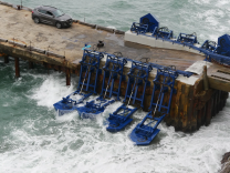 Blue paddle-like devices immersed in ocean surf are attached to a wooden pier. An SUV is parked on the pier.
