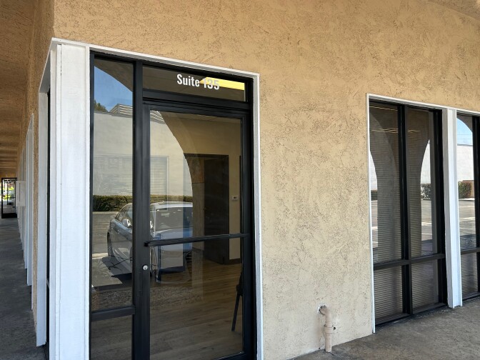An office door in an outdoor strip mall is shown. The glass door does not have signage on it. Above the door it says suite 135. 